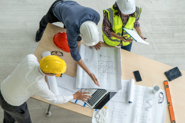 High-angle photo from directly above of an architect, a businessman, and a construction worker examining a blueprint on a construction site.